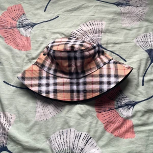 Rutig bucket hat i beige och svart - Snygg bucket hat med klassiskt rutigt mönster i beige, svart, vitt och röda detaljer. Hatten har bred brätte och är perfekt för att lyfta din stil. Tillverkad i mjukt bomullstyg som känns skönt mot huvudet.