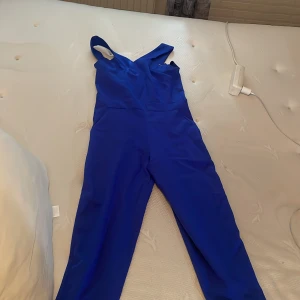 Kornblå byxdress med breda axelband - Snygg kornblå byxdress med breda axelband och v-ringad överdel. Byxdressen har raka ben och är tillverkad i ett slätt, vävt tyg som känns bekvämt och stilrent. Perfekt för dig som vill sticka ut med färg och enkel design.