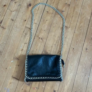 Svart axelväska med kedja från Tiamo - Snygg svart axelväska från Tiamo i skinnimitation med grov silverfärgad kedja som axelrem och runt kanterna. Väskan har dragkedja både på baksidan och inuti, samt flera fack för småprylar. Perfekt storlek för det viktigaste och ger en cool vibe till din outfit. Nypris 550 och lknappt andvönd