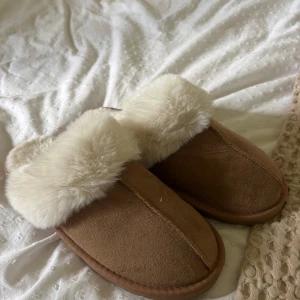Bruna tofflor med fuskpäls från 157 - Mysiga bruna tofflor från 157 med fluffig vit fuskpäls runt öppningen. Utsidan är i mockaliknande material och insidan är supermjuk, perfekta för kalla dagar hemma. Klassisk slip-in modell med rund tå och platt sula.
