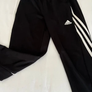 Svarta Adidas träningsbyxor med ränder - Svarta träningsbyxor från Adidas med klassiska vita ränder längs sidorna och Adidas-logga på låret. Byxorna har elastisk midja och är tillverkade i ett lätt och sportigt material som andas. Perfekta för dig som gillar en sportig och clean stil.