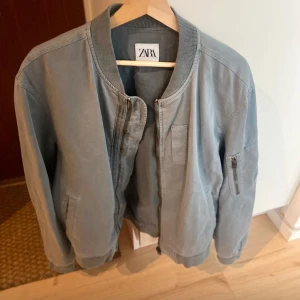 Grå bomberjacka från Zara - Snygg grå bomberjacka från Zara i storlek L. Jackan har klassisk ribbad krage, muddar och dragkedja framtill. Fickor på sidorna och en extra ficka med dragkedja på ärmen. Perfekt för dig som gillar en clean och stilren look.