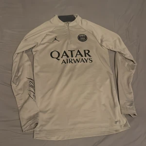 Beige PSG x Jordan half zip tröja - Säljer en beige långärmad PSG x Jordan träningströja med svart tryck och logga på bröstet, samt signaturdetalj på ärmen. Tröjan har half zip och är i slim fit-modell, perfekt för fotbollsträning eller chill. Materialet är polyester.