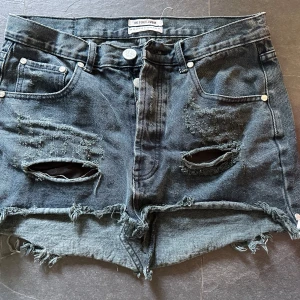 Svart jeanskjol från One Teaspoon - Cool svart jeanskjol från One Teaspoon med rå kant och slitna detaljer framtill. Kjolen har klassiska fickor, knappstängning och en kort passform som ger en edgy vibe. Perfekt för dig som gillar streetstyle och vill sticka ut. Low waste /lågmidjad. Lite längre där bak så rumpan inte syns