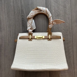 Vit handväska med croco-mönster - Snygg vit handväska i croco-mönstrat syntetmaterial med beige detaljer och guldfärgade metalldelar. Väskan har både handtag och avtagbar axelrem samt dragkedja upptill. Handtaget är dekorerat med en beige och vit scarf med hjärtmönster för extra stilpoäng. Väskan är helt ny och oanvänd🥰
