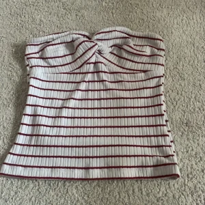 Randig bandeau tubtopp från NA-KD - Superfin bandeau tubtopp från NA-KD i vitt med röda ränder. Toppen är ribbad och har en twistad detalj framtill som ger en snygg touch. Perfekt för varma dagar och enkel att styla med jeans eller kjol.