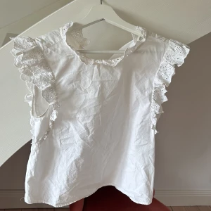 Vit blus med volangärmar XS - Supersöt vit blus i bomull med broderade volangdetaljer längs ärmhålen och halsringningen. Ärmlös modell med romantisk känsla och fina hålbroderier som ger en extra touch. Perfekt för dig som gillar en feminin och luftig stil💞