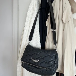  Zadig & Voltaire - Cool svart axelväska i quiltat skinn från Zadig & Voltaire med stjärnmönster, bred justerbar axelrem och silverfärgade metalldetaljer. Väskan har dragkedja både fram och bak samt ikonisk vinglogga framtill. Perfekt för att lyfta din outfit.