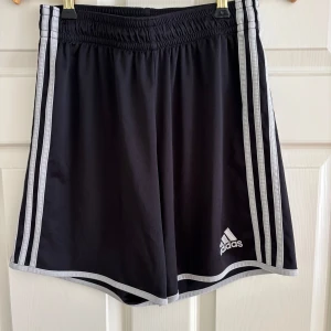 Svarta Adidas shorts med vita ränder - Klassiska svarta Adidas shorts med vita ränder längs sidorna och vit logga framtill. Elastisk midja och sportig passform, perfekta för träning eller chill. Lätta och bekväma i syntetmaterial.