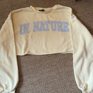 Gul croppad sweatshirt IN NATURE - Croppad sweatshirt i ljusgul färg från med trycket 'IN NATURE' i stora ljuslila bokstäver över bröstet. Tröjan har långa ärmar, rund halsringning och är gjord i mjuk bomullsmix. Perfekt för en chill och trendig look.
