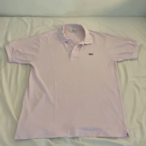 Ljusrosa pikétröja från Lacoste - Säljer en ljusrosa pikétröja från Lacoste med klassisk krage och knäppning framtill. Tröjan har det ikoniska gröna krokodilmärket broderat på bröstet. Tillverkad i bomull och har korta ärmar. Perfekt för en clean och sportig stil.