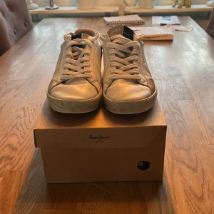 Vita och svarta sneakers från Pepe Jeans - Snygga sneakers från Pepe Jeans i vitt och svart med grå detaljer. Klassisk låg modell med rund tå, snörning och platt sula. Ovandel i skinn och textil, diskret logga på sidan och årtalet 1973 på hälen. Perfekta för en stilren och avslappnad look. Storlek 41 Skriv vid frågor 