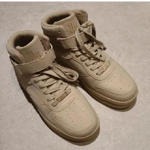 Beiga skor från Bershka - Snygga beige sneakers från Bershka med hög modell, snörning och band i stlk 41. Skorna har en bred rem över vristen för extra stil och komfort. Perfekta för en trendig look. Endast testade.