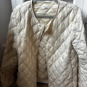 Quiltad beige jacka från Uniqlo - Quiltad beige jacka från Uniqlo med rund halsringning och tryckknappar framtill. Jackan är lätt vadderad och har ett stilrent rutmönster. Perfekt för lager-på-lager och enkel att matcha med olika outfits.