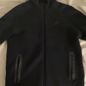 Svart Nike Tech - Svart vindjacka från Nike med stilren design och diskret logga på bröstet. Jackan har hel dragkedja framtill och två fickor med dragkedja. Perfekt för dig som gillar sportig och clean stil.
