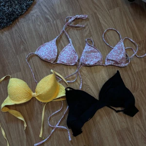 Fyra  snygga bikinis i olika färger - Säljer tre bikinis: en vit med rosa blommigt mönster och smala knytband, en gul med vadderade kupor och spetsdetaljer, samt en enkel svart bikini med triangelformade kupor. Perfekt för stranden eller poolen i sommar! Alla har klassisk bikini-modell. Varje bikini överdel kostar 40kr st 