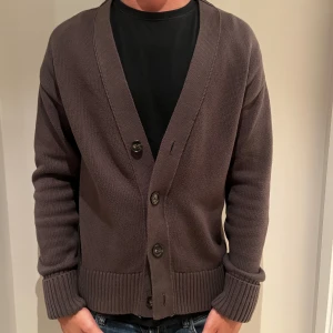 Brun stickad kofta med knappar från John Henric - Säljer en brun stickad kofta med djup v-ringning och stora knappar framtill. Koftan har långa ärmar och breda ribbade muddar vid ärmslut och nederkant. Perfekt för lager-på-lager och chill stil. Nästan som ny, passar S/M 