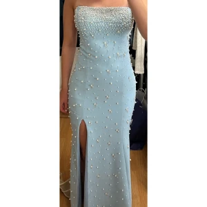 Ljusblå långklänning med pärlor - Ljusblå festklänning från MK Mode i Södertälje Figurnära klänning med glittrande pärldekor över hela plagget, förutom på baksidan. Modellen har en slits framtill och en axelbandslös design, smala axelband finns om man vill sy på dem. Matchande puffärmar i satinliknande material kan också fästas på.  Passar XS/S. Perfekt till bröllopsfest eller andra festliga tillfällen. Köpt för 4 500 kr och endast använd en kväll, i mycket bra skick.