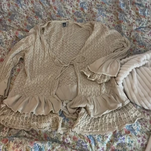 Beige kofta med spets och volanger - Superfin beige kofta med spetsdetaljer och volanger längs nederkant och ärmslut. Koftan har ett snyggt stickat mönster och knytband framtill. Perfekt för dig som gillar romantisk och boho stil. Långärmad och riktigt mysig att ha på sig.