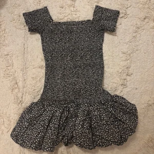 Svart blommig kortklänning - Supersöt svart kortklänning med små vita blommor och volang nertill. Klänningen har korta ärmar och en smickrande, figurnära passform. Perfekt för dig som gillar en feminin och trendig stil.