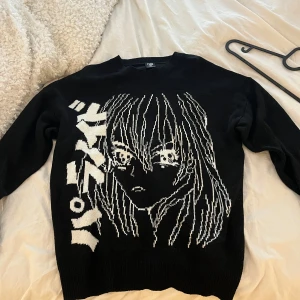 Svart stickad anime-tröja från FSBN - Svart stickad tröja från FSBN med relaxed fit och stort vitt anime-motiv framtill samt japansk text. Tröjan har rund halsringning och långa ärmar. Perfekt för dig som gillar streetstyle och manga vibes.
