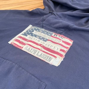 Mörkblå hoodie Ralph Lauren Denim & Supply - Snygg mörkblå hoodie från Ralph Lauren Denim & Supply med stor patch framtill i amerikansk flaggdesign och text. Tröjan har huva med snörning och klassisk känguruficka. Materialet är mjuk bomull, perfekt för chill dagar. Eller när du vill sticka ut på stan. 
