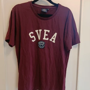 Vinröd t-shirt från SVEA - Snygg vinröd t-shirt från SVEA med vit logotyp och emblem på bröstet. Klassisk rund hals och korta ärmar. Perfekt för dig som gillar streetwear och enkel stil. Tillverkad i mjuk bomull som är skön mot huden.