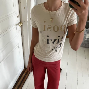 Beige t-shirt med tryck från Name It - Snygg beige t-shirt från Name It med coolt tryck i svart och guld där det står 'positivity'. Modellen är klassisk med rund hals och korta ärmar. Mjuk bomullskvalitet som är skön mot huden. Perfekt för dig som gillar statement-plagg och stilrena färger.