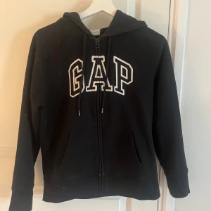 Svart hoodie från GAP - Säljer en klassisk svart hoodie från GAP med dragkedja och stor logga på bröstet. Tröjan har långa ärmar och en praktisk huva. Perfekt för en avslappnad stil.