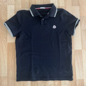 Svart pikétröja från Moncler - Snygg svart pikétröja från Moncler med klassisk krage och knappar. Tröjan har vita detaljer på kragen och ärmsluten samt Monclers logga broderad på bröstet. Perfekt för en stilren look.