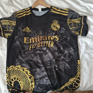 Svart och guld fotbollströja från Adidas - Snygg svart fotbollströja med guldiga detaljer och drakmönster. Tröjan har korta ärmar och klubbens emblem på bröstet. Perfekt för fotbollsfans som vill sticka ut på planen.
