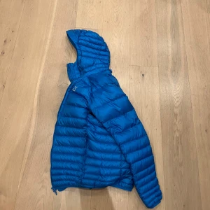 Blå dunjacka från Haglöfs - Säljer en snygg blå dunjacka från Haglöfs. Jackan har en quiltad design och är utrustad med en praktisk huva. Perfekt för kyligare dagar och håller dig varm och bekväm. Passar bra för både vardag och utomhusaktiviteter. Ända defekten är lagningen på ena armen. 