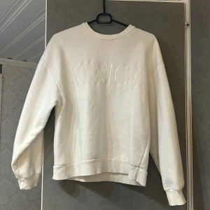 Vit sweatshirt med texten 'VENICE' - Säljer en stilren vit sweatshirt med upphöjd text 'VENICE' på framsidan. Tröjan har långa ärmar och ribbade muddar vid ärmslut och nederkant. Perfekt för en avslappnad stil.