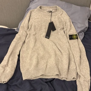 Grå stickad tröja från Stone Island - Säljer en stilren grå stickad tröja från Stone Island. Tröjan har en rund halsringning och långa ärmar med ribbade muddar. Perfekt för kyligare dagar. Märkets ikoniska logga finns på ärmen. Den är 14-16 men passar mig som är 172 och 58 kg, skulle säga att den passer från 168-175