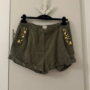 Olivgröna shorts med dekorativa stenar - Snygga olivgröna shorts från H&M med glittrande stenar på fickorna. Perfekta för en somrig look med en touch av glamour. Shortsen har en bekväm passform och är gjorda i ett mjukt material.