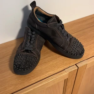 Gråa Louboutins med bitar - Snygga gråa Louboutins med nitar på tån och svarta snören. Skorna har en unik design med röd sula och är perfekta för en som söker efter pappas pengar stilen. Finns ett litet hål i hälen på högra skon men det märks inte av så länge man inte springer och hoppar. Storlek 43 men passar 44 om man har lite smalare fötter.