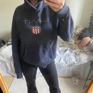 Mörkblå hoodie från Gant - Säljer en snygg mörkblå hoodie från Gant med deras klassiska logga och emblem på bröstet. Tröjan har en stor ficka framtill och en bekväm huva. Perfekt för en avslappnad stil.