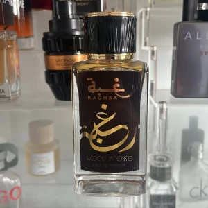 Raghba Wood Intense Eau de Parfum - Säljer en elegant flaska Raghba Wood Intense Eau de Parfum. Flaskan är rektangulär med en svart och guld etikett och har en svart kork med gulddetaljer. Parfymen är tillverkad i U.A.E och har en rik, träig doftprofil.