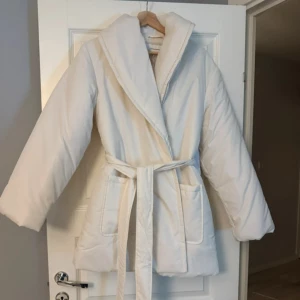 Vit vadderad jacka från Bubbleroom - Säljer en stilren vit vadderad jacka från Bubbleroom. Jackan har ett bälte i midjan och långa ärmar, perfekt för kyliga dagar. Den är designad för att hålla dig varm och bekväm med sin mjuka vaddering.