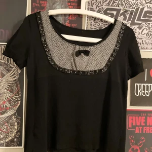 Svart kortärmad topp med dekorativ krage - Snygg svart topp med korta ärmar och en unik krage i grått mönster. Kragen är dekorerad med svarta pärlor och en söt rosett. Köpt second hand vet inte vilket märke (står inget)😊 pris går att diskutera