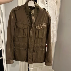 Olivgrön jacka från Moncler -  En Grön Fieldjacka från Moncler, jackan är en size 3 vilket motsvarar ungefär en M. Jackan är producerad under tidigt 2000-tal (därav brun label) och är införskaffad på Garmsmarket som garanterar äkthet. Den är inte fodrad och passar därför vår sommar bäst. Jackan är i 7/10 cond och tvättas självklart innan frakt. 
