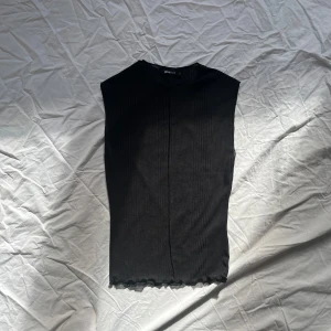 Svart ribbad ärmlös topp från Gina Tricot - Snygg svart ribbad topp från Gina Tricot. Den är ärmlös och har en enkel design med en liten vågig kant vid nederdelen. 