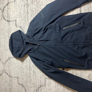 Mörkblå vindjacka från Woolrich - Säljer en stilren mörkblå vindjacka från Woolrich. Jackan har en praktisk huva och dragkedja framtill. Två fickor med dragkedja på sidorna ger extra funktionalitet. Perfekt för blåsiga dagar. Ny pris 3500
