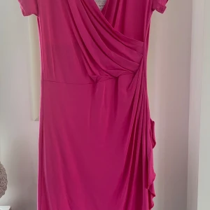 Rosa omlottklänning från NOT - Snygg rosa omlottklänning från NOT i mjukt och stretchigt material. Klänningen har korta ärmar och en v-ringad design med draperade detaljer på sidan. Perfekt för en färgglad look!