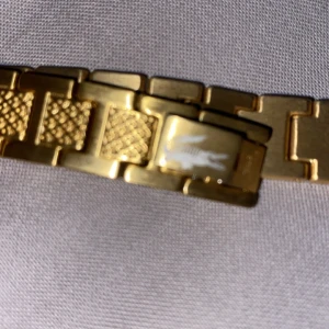 Lacoste armband  - Elegant guldarmband med ett unikt krokodilmotiv. Armbandet har en stilren design med texturerade länkar som ger det en exklusiv känsla. Perfekt för att ge en touch av lyx till din stil.