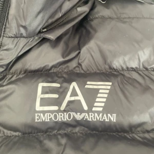 Svart dunjacka från Emporio Armani - Snygg svart dunjacka från Emporio Armani EA7 med dragkedja och huva. Perfekt för kyliga dagar med stilren design och logotyp på bröstet, med tanke på att den har blivit för stor för mig. Haft den i 2 år men endast använt 5 gånger så jackan är alltså ny kan man säga. Inga repor eller liknande utan är nyskick. Är liten i storlek så jag rekomenderar dem som har storlek L att köpa denna jackan! Köptes för 1800 kr och säljer nu för ett billigare pris.