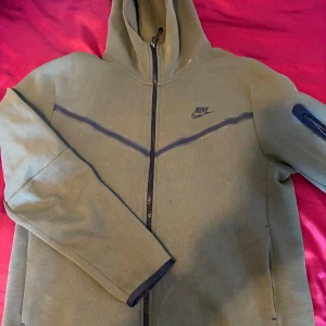 Nike tech flees grön - Säljer en olivgrön hoodie från Nike med dragkedja och huva. Jackan har en praktisk ficka med dragkedja på ärmen och svarta detaljer. Perfekt för en sportig look! Har även defekter (se bild 5), det är ett litet hål på insidan som INTE leder till ytterficka. Det är alltså 2 helt olika fickor!  Tvättas såklart innan den postas❤️