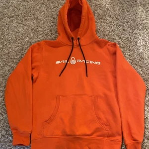Orange hoodie från Sail Racing - Säljer en snygg orange hoodie från Sail Racing med vit logga på bröstet. Tröjan har en klassisk känguruficka och justerbar huva med svarta snören. Perfekt för en avslappnad stil.