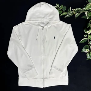 Vit hoodie från Ralph Lauren - Säljer en stilren vit hoodie från Ralph Lauren med dragkedja och det klassiska logotypen på bröstet. Tröjan har långa ärmar och en praktisk huva. Perfekt för en avslappnad look.