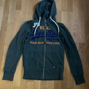 Grön hoodie från Superdry - Säljer en grön hoodie från Superdry, i storlek XS med dragkedja och tryck i blått och orange på framsidan. Den har en justerbar huva med vita snören och långa ärmar. Perfekt för en avslappnad stil. 